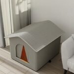 Graues isoliertes Katzenhaus mit oranger Türklappe in einem hellen Wohnzimmer neben einem Sessel