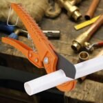 Oranger Ratschen-Rohrschneider schneidet ein weißes Kunststoffrohr auf einer Werkbank mit Messingfittings