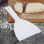 Weißer Teigschaber aus Kunststoff auf einer bemehlten Arbeitsfläche vor einem Holzbrett mit Teig